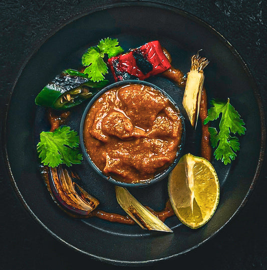 A black plate featuring a bowl of roasted salsa tatemada surrounded by grilled vegetables, including charred red and green peppers, caramelized onions, lime wedges, and fresh cilantro leaves.