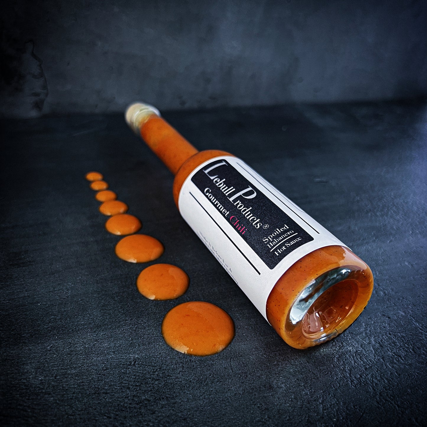 Bottle of Spoiled Habanero hot sauce lying on a dark textured surface with drops of orange sauce arranged in a line, showcasing its smooth and vibrant consistency.